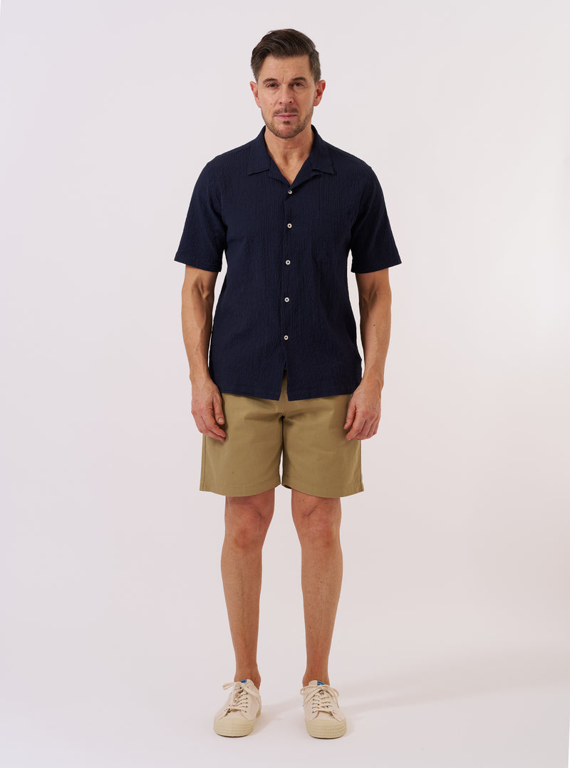 The Navy Seersucker Shirting Road Shirt is shown as a flat lay against a plain white background. The shirt features a camp collar, a central button-down front with tonal buttons, and a single chest patch pocket on the left side. The fabric has a distinct puckered seersucker texture in a deep navy blue.