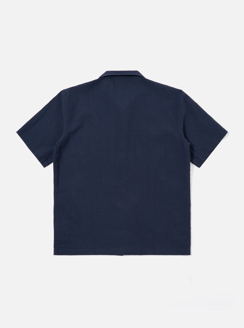 The Navy Seersucker Shirting Road Shirt is shown from a rear angle as a flat lay against a plain white background. The image highlights the shirt's short sleeves, straight hem, and the subtle puckered texture of the navy blue seersucker fabric. A small locker loop is visible at the center of the back yoke.