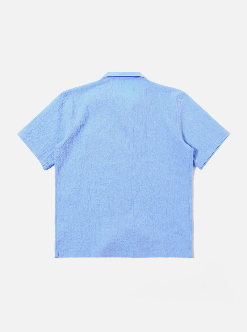 The Sky Blue Seersucker Shirting Road Shirt is shown from the back as a flat lay against a plain white background. The image highlights the shirt's short sleeves, straight hem, and the puckered texture of the sky blue seersucker fabric.