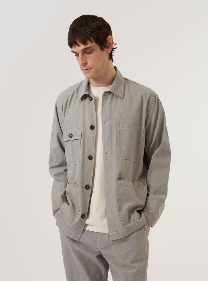 A model wears the Grey Izumi Cotton Dockside overshirt against a plain white background. The overshirt features a button-down front, a classic collar, and two large patch pockets at the waist. The garment is made from a textured grey cotton fabric and has a relaxed fit.