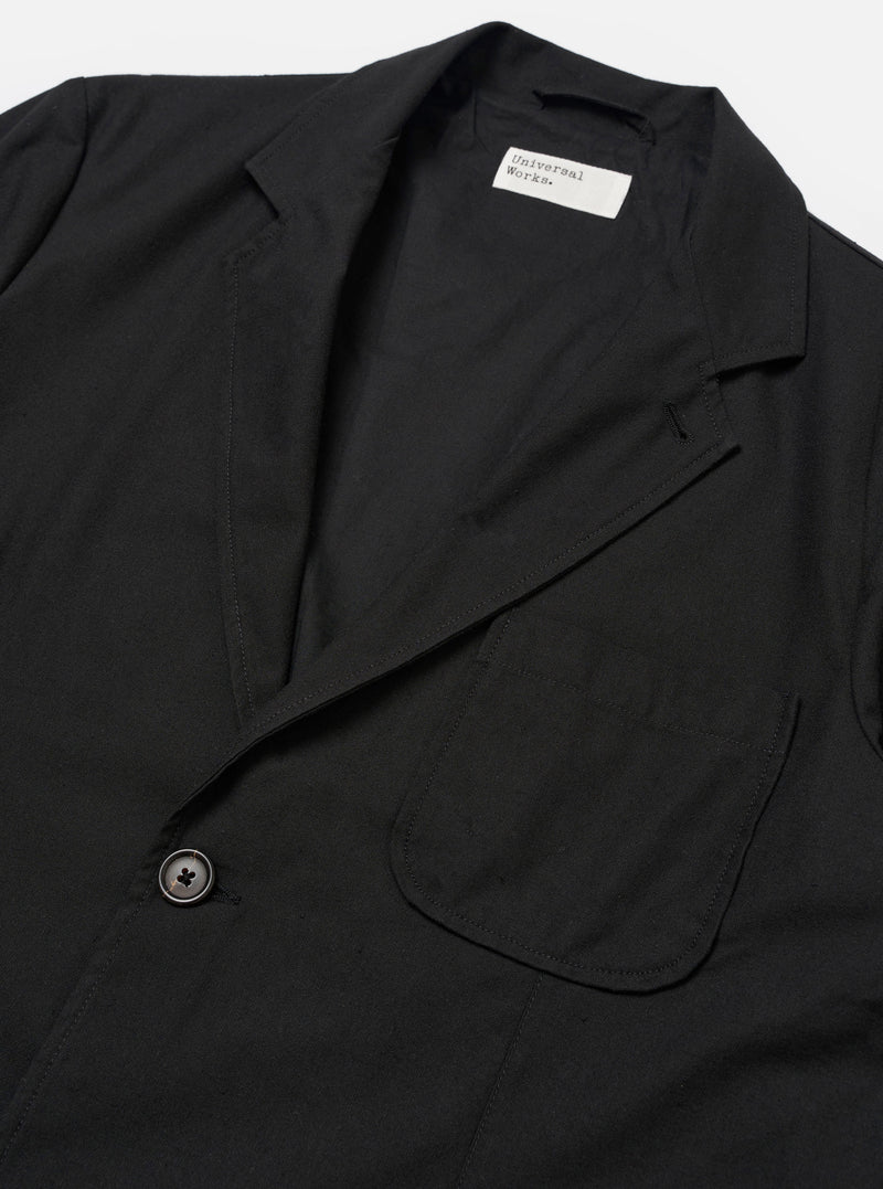 The Black Valencia Cotton Two Button Jacket is worn by a model against a plain white background. The image shows a rear three-quarter view, highlighting the jacket's loose fit, structured shoulders, and central back seam. The black fabric has a smooth finish, and the model is wearing the jacket over a light-colored long-sleeve top. No mannequin is present in the image.