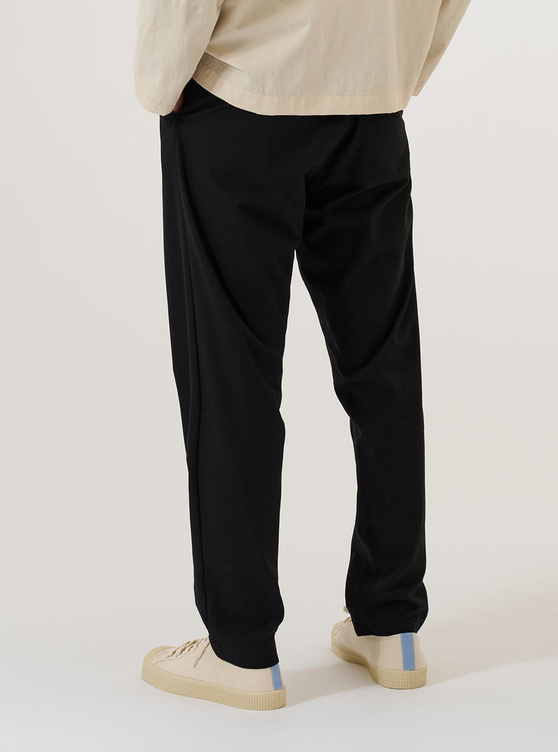 A model wears the Black Valencia Cotton Military Chino, shown from a side profile against a plain white background. The image highlights the straight-leg fit and the clean construction of the black Valencia cotton fabric. The model is wearing the trousers from the waist down, paired with a light-colored top and dark shoes.