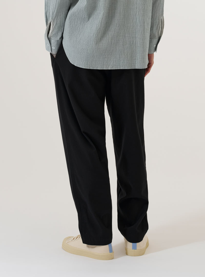 A model wears the Black Valencia Cotton Pleated Track Pant, shown from the waist down against a plain white background. The image highlights the loose, tapered fit of the trousers, the single pleat at the front, and the elasticated waistband with a drawstring. The black cotton fabric has a smooth finish, and the model is wearing the pants with a white t-shirt and white socks.