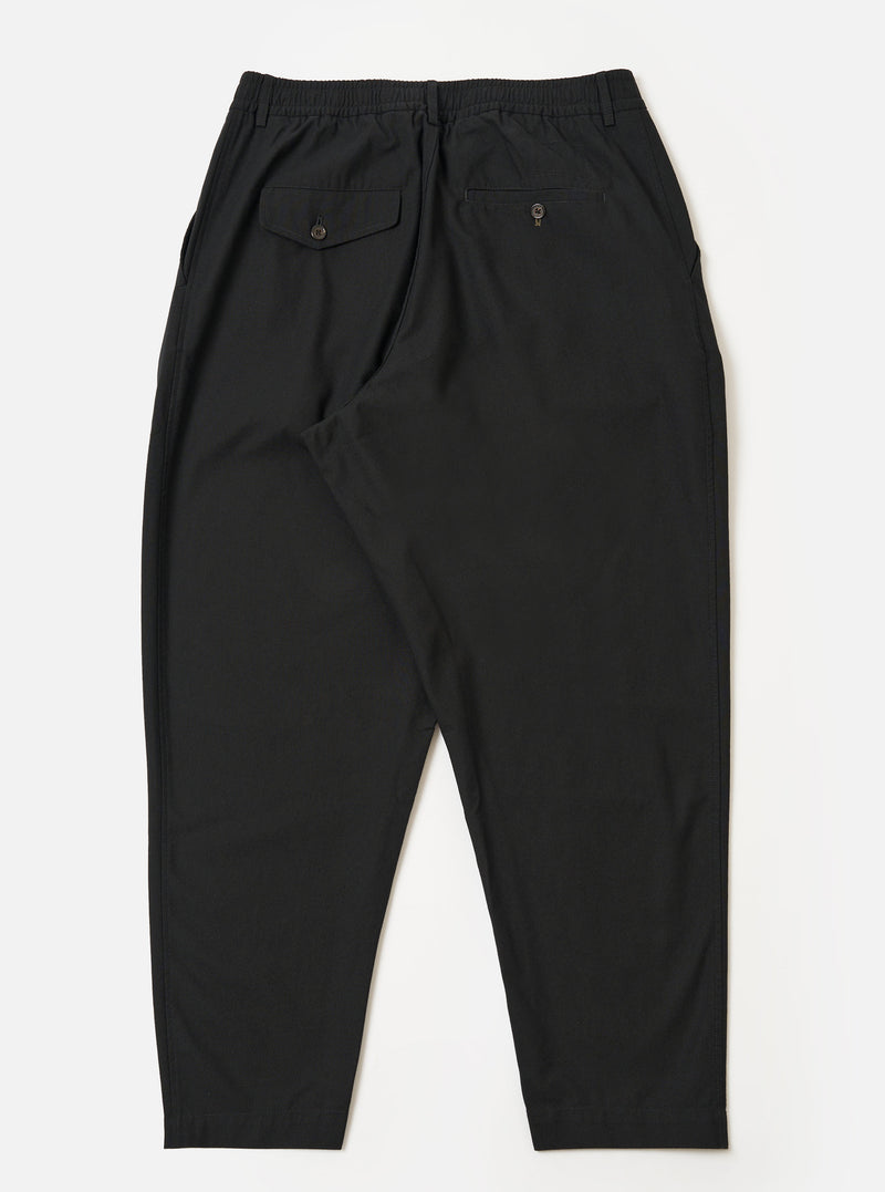 The Black Valencia Cotton Pleated Track Pant is shown from the back as a flat lay against a plain white background. The image highlights the elasticated waistband and the two rear pockets: one featuring a buttoned flap and the other a button jetted design. The black fabric has a clean, fine plain weave texture.