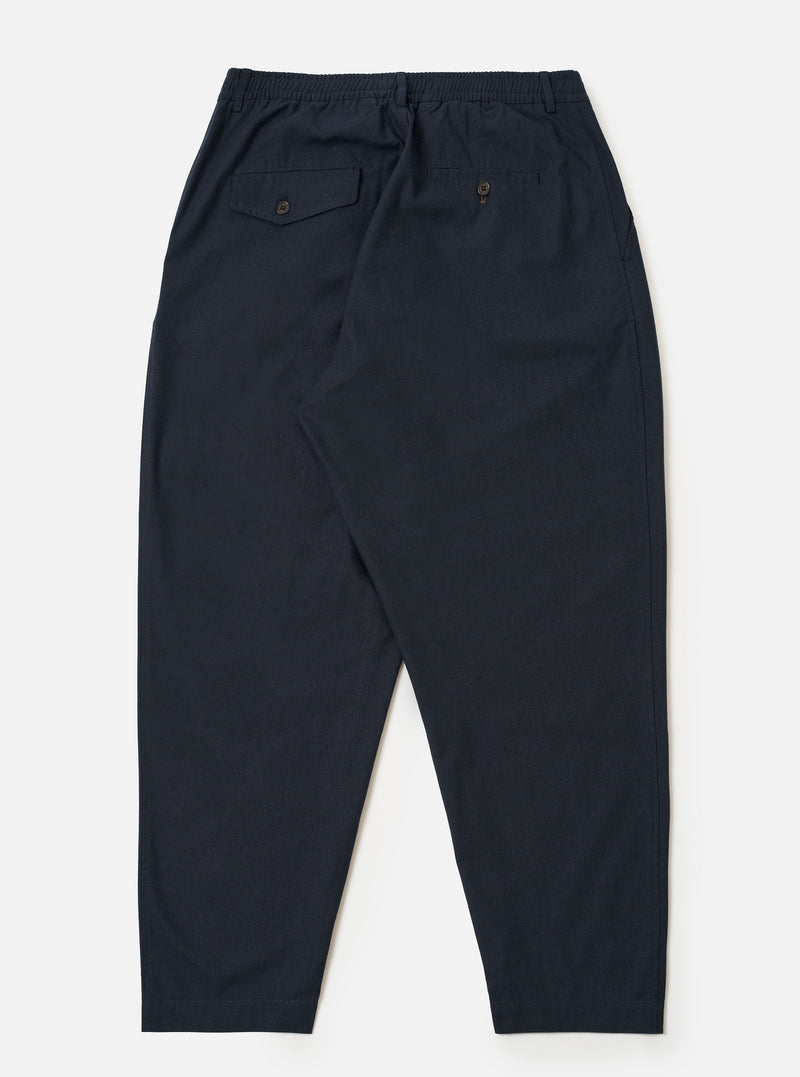 The Navy Valencia Cotton Pleated Track Pant is shown from the back as a flat lay against a plain white background. The image highlights the two rear pockets, including one with a buttoned flap and one button jetted pocket, and the elasticated waistband. The navy blue fabric has a fine, plain weave.