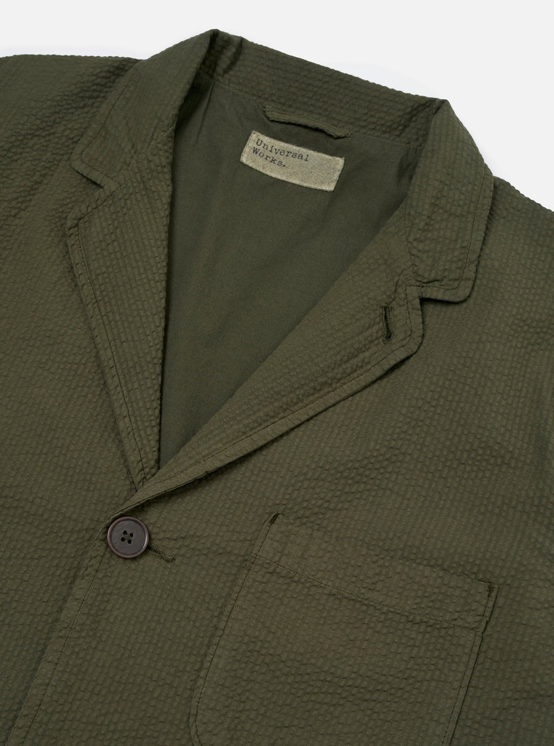 The Olive Merida Seersucker Three Button Jacket is shown as a flat lay against a white background. The jacket features a three-button front closure, a notch lapel, and two large patch pockets at the hips. The olive green fabric has a distinct puckered seersucker texture throughout.