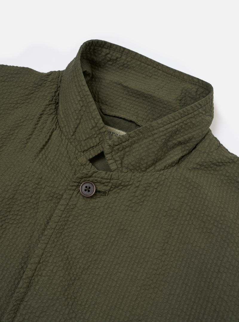 The Olive Merida Seersucker Three Button Jacket is shown as a flat lay against a white background. The jacket features three buttons, a classic lapel, one chest patch pocket, and two lower patch pockets. The olive green cotton fabric has a characteristic puckered seersucker texture.