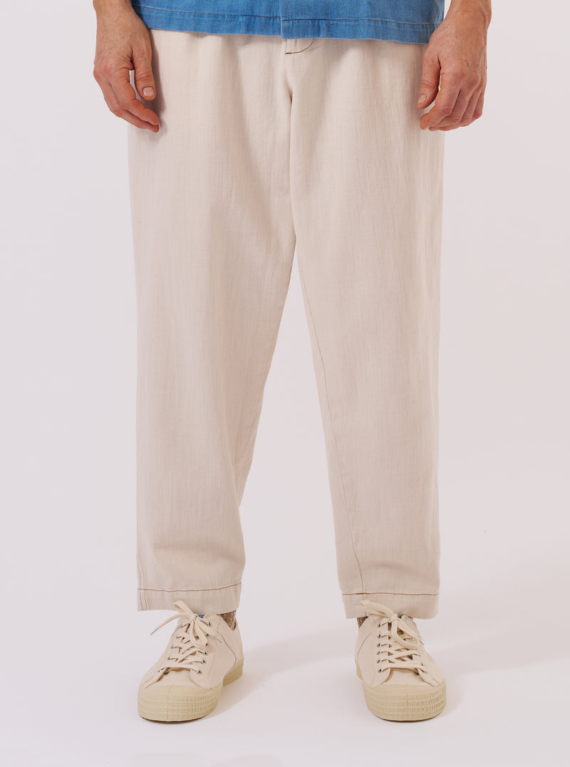 A model wears the Ecru Koko Cotlin Pleated Track Pant against a plain white background. The image shows the model from the waist down, highlighting the loose, tapered fit and the single pleat at the front. The ecru-colored fabric has a visible slubby, textured appearance. The model is also wearing white socks and dark-colored shoes.