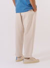 A model wears the Ecru Koko Cotlin Pleated Track Pant, shown from the back against a plain white background. The image highlights the relaxed, tapered fit of the trousers and the single rear patch pocket with a buttoned flap on the right side. The ecru-colored cotton-linen blend fabric has a visible slubby, textured appearance. The model is wearing the trousers with white socks and dark shoes.