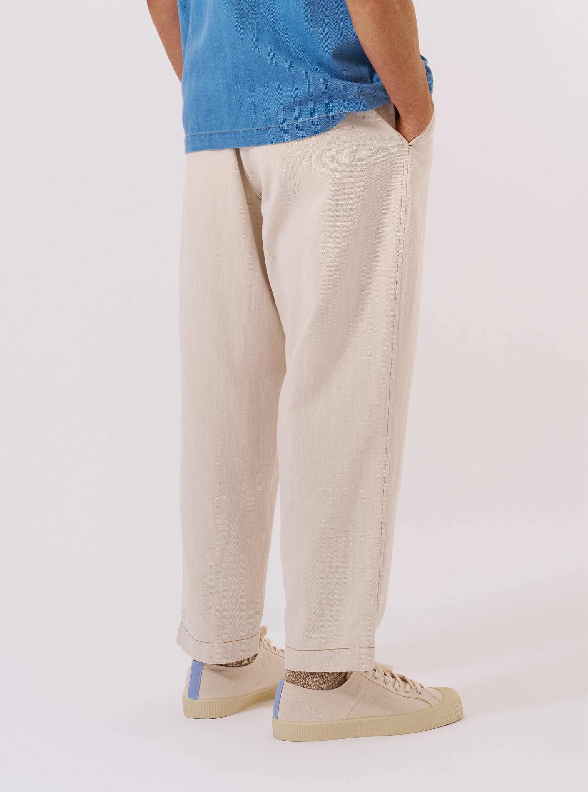 A model wears the Ecru Koko Cotlin Pleated Track Pant, shown from the back against a plain white background. The image highlights the relaxed, tapered fit of the trousers and the single rear patch pocket with a buttoned flap on the right side. The ecru-colored cotton-linen blend fabric has a visible slubby, textured appearance. The model is wearing the trousers with white socks and dark shoes.