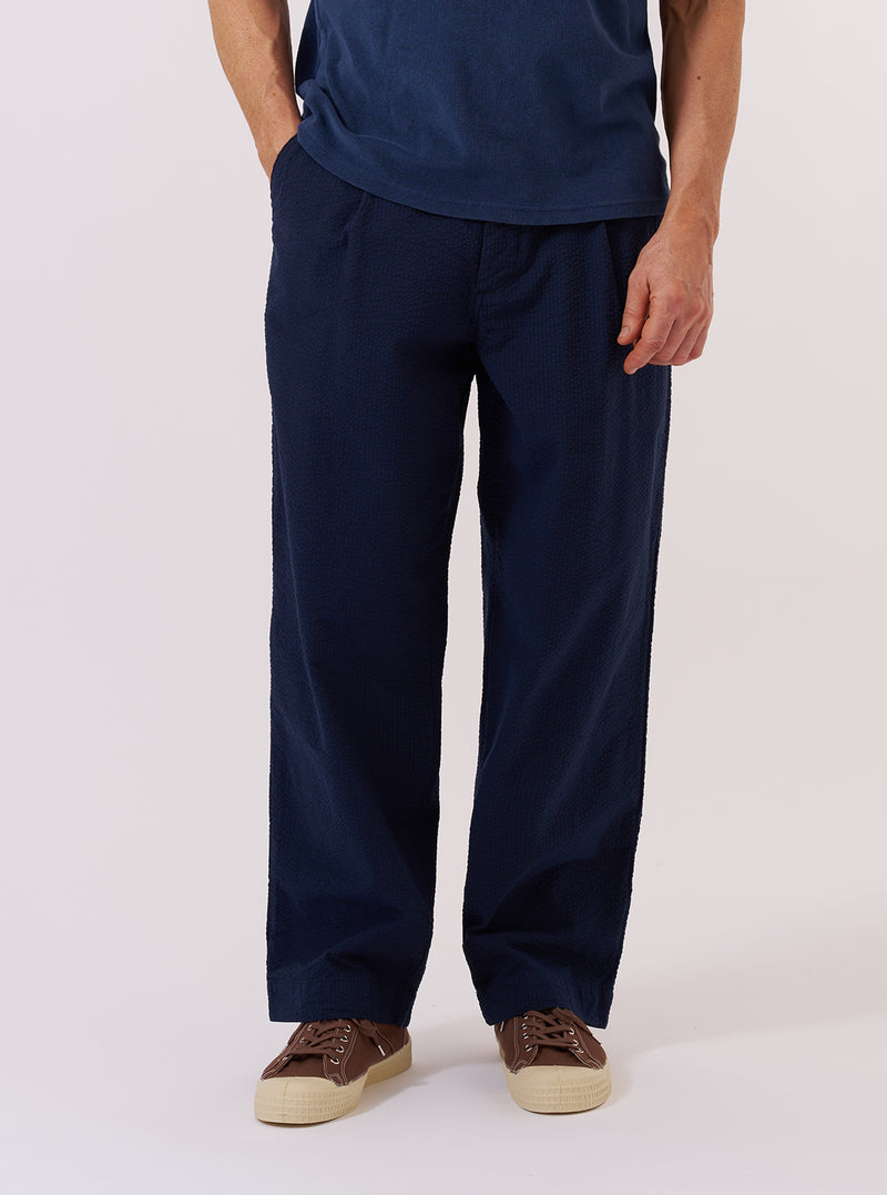 A model wears the Navy Derby Stripe Duke Pant against a plain, neutral background. The image highlights the trousers' loose, wide-leg fit, front pleats, and the vertical navy blue stripe pattern. The model is shown from the waist down, demonstrating how the fabric drapes and the positioning of the waistband.