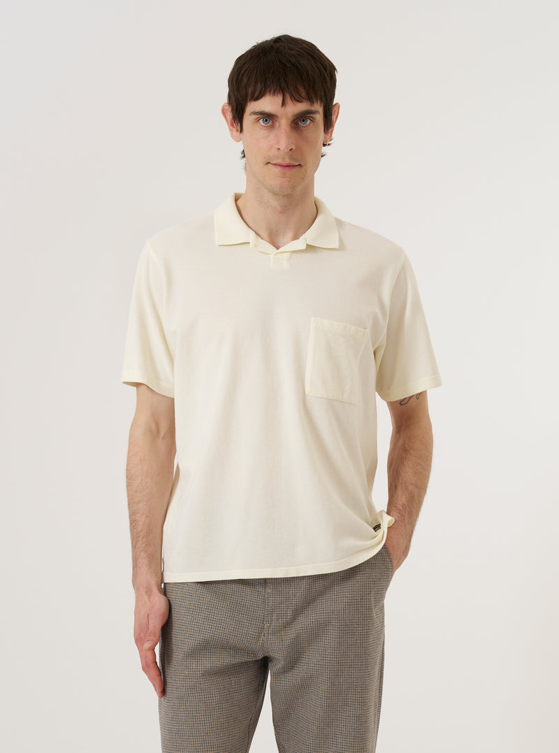 A model wears the Ecru Piquet Vacation Polo against a plain white background. The polo features a buttonless open neck, a single-piece collar, and a single patch pocket on the left chest. The ecru-colored cotton piquet fabric has a visible fine honeycomb texture. The model is shown from the chest up, highlighting the relaxed fit and clean design of the neckline.