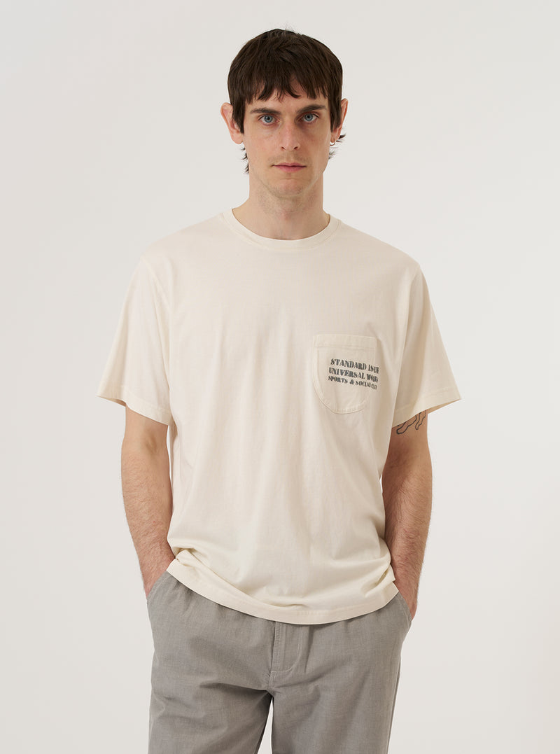 A model wears the Ecru Organic Jersey Print Pocket Tee against a plain, neutral background. The short-sleeved t-shirt features a chest pocket with a printed design and is made from a soft ecru-colored organic jersey fabric.
