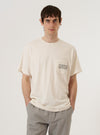 A model wears the Ecru Organic Jersey Print Pocket Tee against a plain, neutral background. The short-sleeved t-shirt features a chest pocket with a printed design and is made from a soft ecru-colored organic jersey fabric.