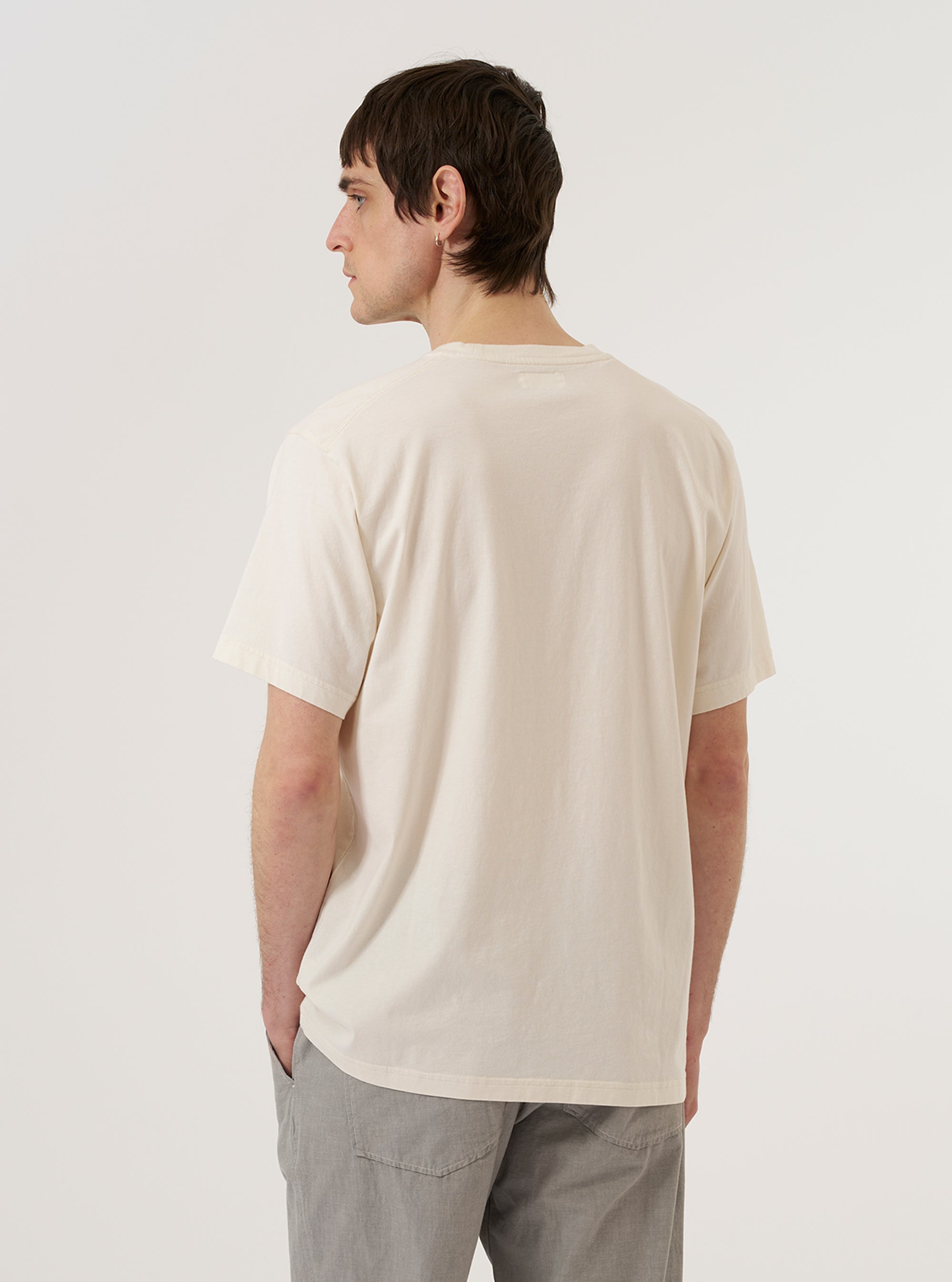 A model wears the Ecru Organic Jersey Print Pocket Tee, shown from the back against a plain white background. The t-shirt features a relaxed fit, short sleeves, and a ribbed crew neckline. The ecru-colored organic cotton jersey fabric has a smooth, clean appearance.