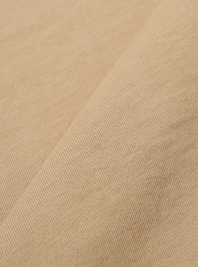 A close-up fabric swatch of the Sand Rugby Jersey Oversized Vacation Polo highlights the sand-colored organic cotton jersey material and its soft, knit texture. The fabric fills the frame, providing a detailed view of the garment's color.