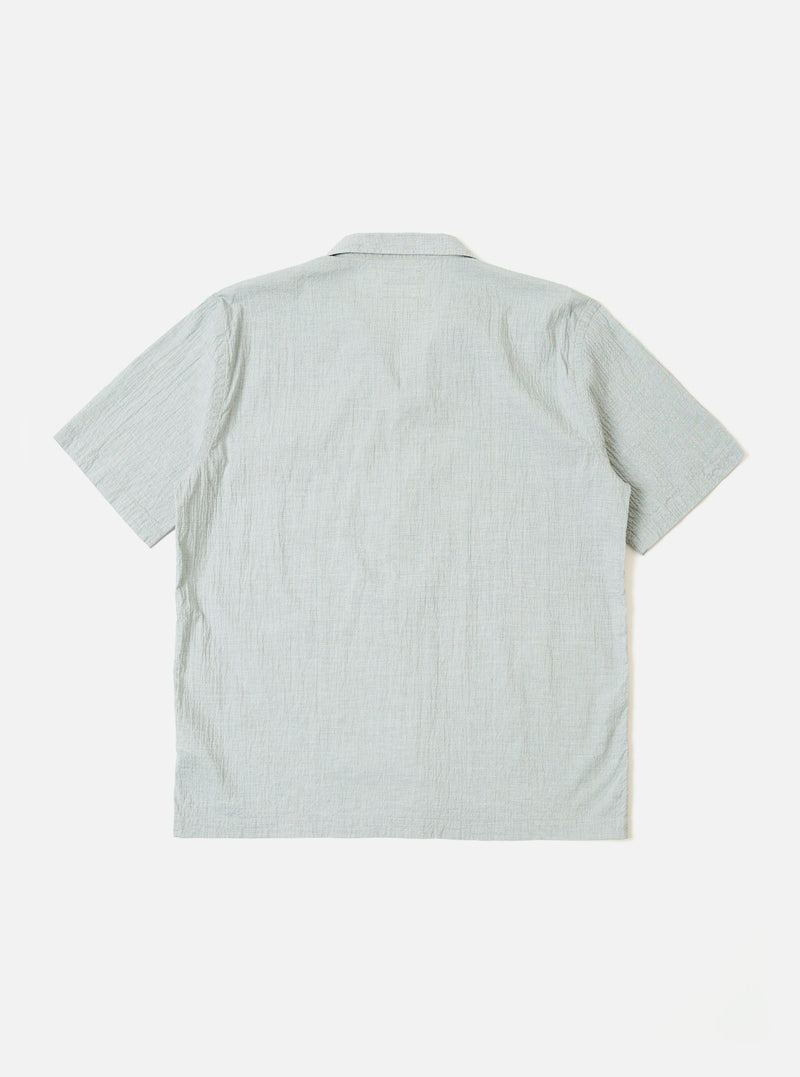 The Green Seersucker Road Shirt is shown from a rear angle on a model. The shirt features a straight hem and short sleeves, with the textured seersucker fabric being prominent. The model is standing against a plain, light grey background. No mannequin is present in the image.