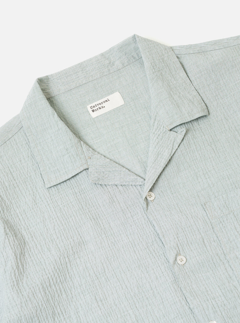The Green Seersucker Road Shirt is worn by a model against a plain, neutral background. The shirt features a relaxed camp collar, a button-front closure, and a single chest pocket. The short-sleeve garment is made from a green cotton fabric with a distinct, puckered seersucker texture. The model is shown from the front, wearing the shirt untucked.