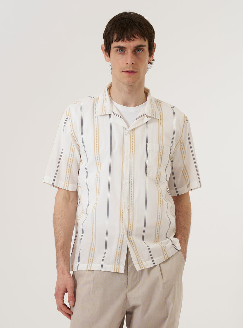 A model wears the Ecru/Navy/Sand Hendrix Stripe Road Shirt against a plain, neutral background. The shirt features a camp collar, a central button-down closure, and vertical stripes in navy, sand, and ecru. The model is shown from the front, highlighting the relaxed fit and the textured stripe pattern of the cotton fabric.