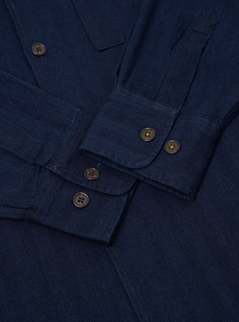 The Indigo Recycled Herringbone Attendant Shirt is shown from the back as a flat lay against a white background. The shirt features a straight hem, a single horizontal yoke across the shoulders, and a simple flat collar. The indigo fabric has a visible vertical herringbone texture throughout.