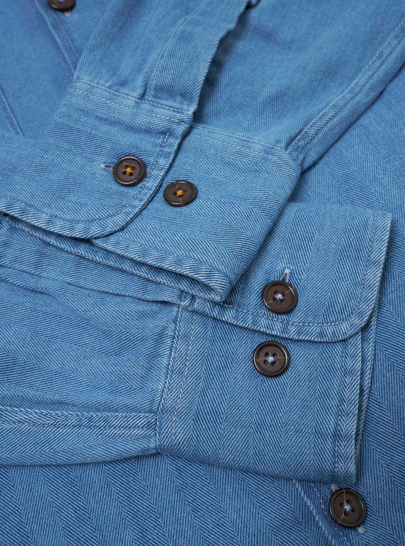 The Washed Indigo Recycled Herringbone Attendant Shirt is shown from the back as a flat lay against a plain white background. The image highlights the clean construction of the shirt's rear panel and the distinct V-shaped texture of the washed indigo herringbone fabric. The garment features a simple straight hem and a yoke across the shoulders.