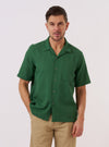 A model wears the Green Letts Cotton Road Shirt over a white t-shirt, standing against a plain white background. The shirt features a camp collar, a central button-down closure, and two large chest patch pockets. The green cotton fabric has a visible textured weave.