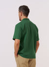 A model wears the Green Letts Cotton Road Shirt over a light-colored top, shown from a rear three-quarter angle against a plain white background. The shirt features a camp collar, a straight hem, and a relaxed silhouette. The olive green cotton fabric has a soft, textured appearance.