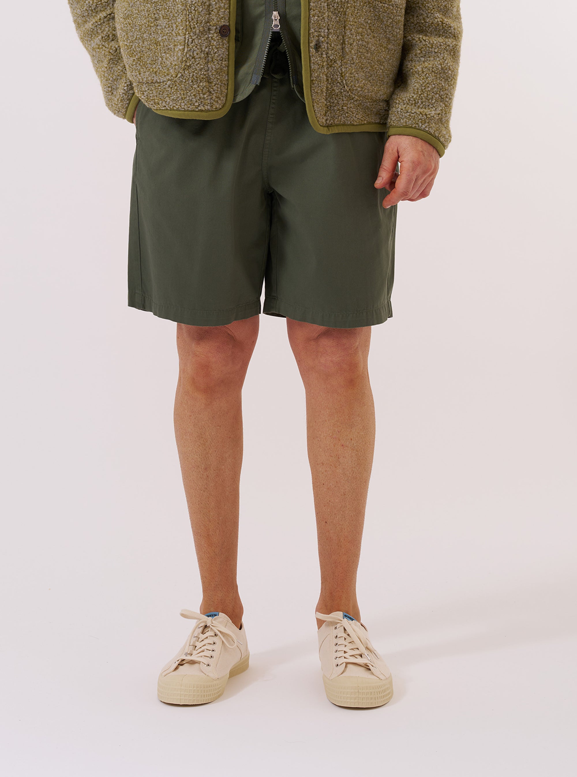 A model wears the Olive Kelly Cotton Simple Short while standing against a plain white background. The shorts feature a relaxed fit with an elasticated waistband and side pockets. The image highlights the soft texture and olive green color of the cotton fabric.