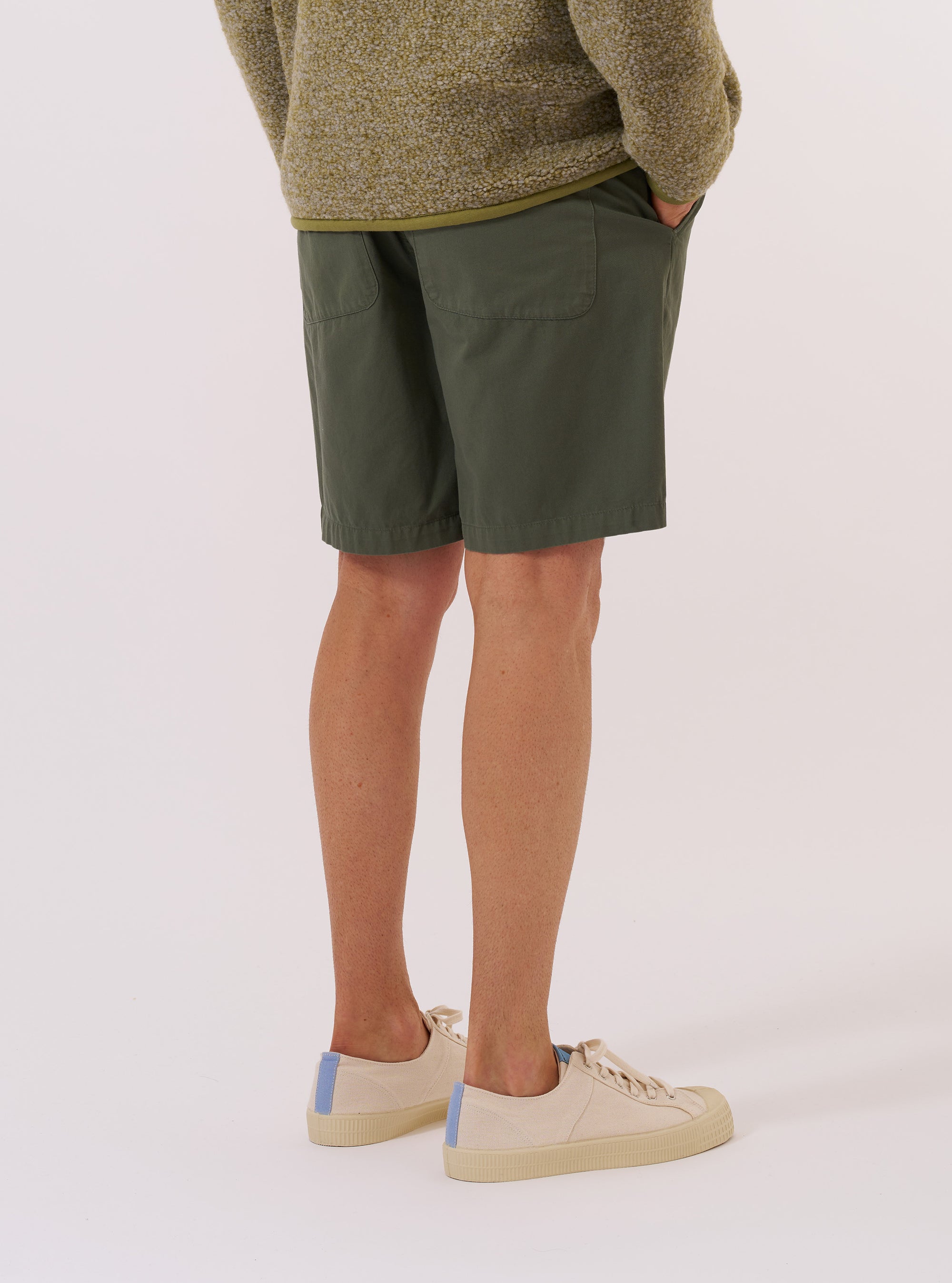 A model wears the Olive Kelly Cotton Simple Short, shown from a rear three-quarter angle against a plain white background. The shorts feature a relaxed fit, an elasticated waistband, and a single patch pocket on the right rear. The olive green cotton fabric has a smooth, matte finish.