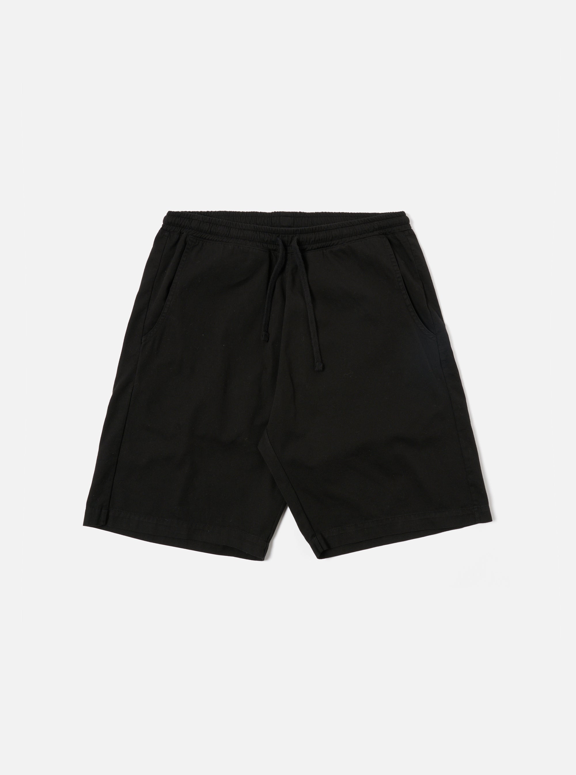 The Black Kelly Cotton Simple Short is shown as a flat lay against a white background. The shorts feature an elasticated waistband with a drawstring, two slanted side pockets, and a single rear patch pocket. The fabric is a solid black cotton with a slight texture.