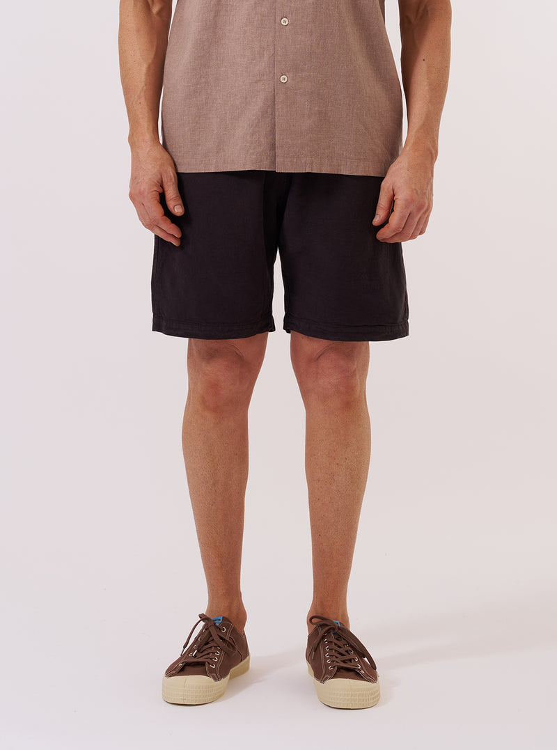 A model wears the Licorice Lyo Linen Mix Simple Short against a plain white background. The shorts feature a relaxed fit, an elasticated waistband with a drawstring, and side pockets. The fabric has a textured, lightweight appearance in a dark brown licorice color. The model is shown from the waist down, paired with a light-colored shirt and dark sandals.