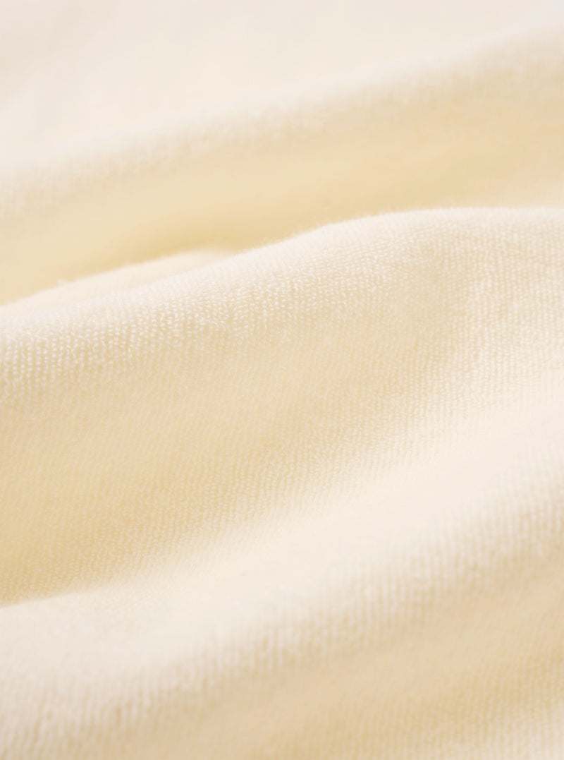 A close-up fabric swatch of the Ecru Light Weight Terry Newlyn Polo highlights the off-white color and the soft, looped texture of the lightweight terry cloth. The material fills the frame, providing a detailed view of the garment's pile and construction.