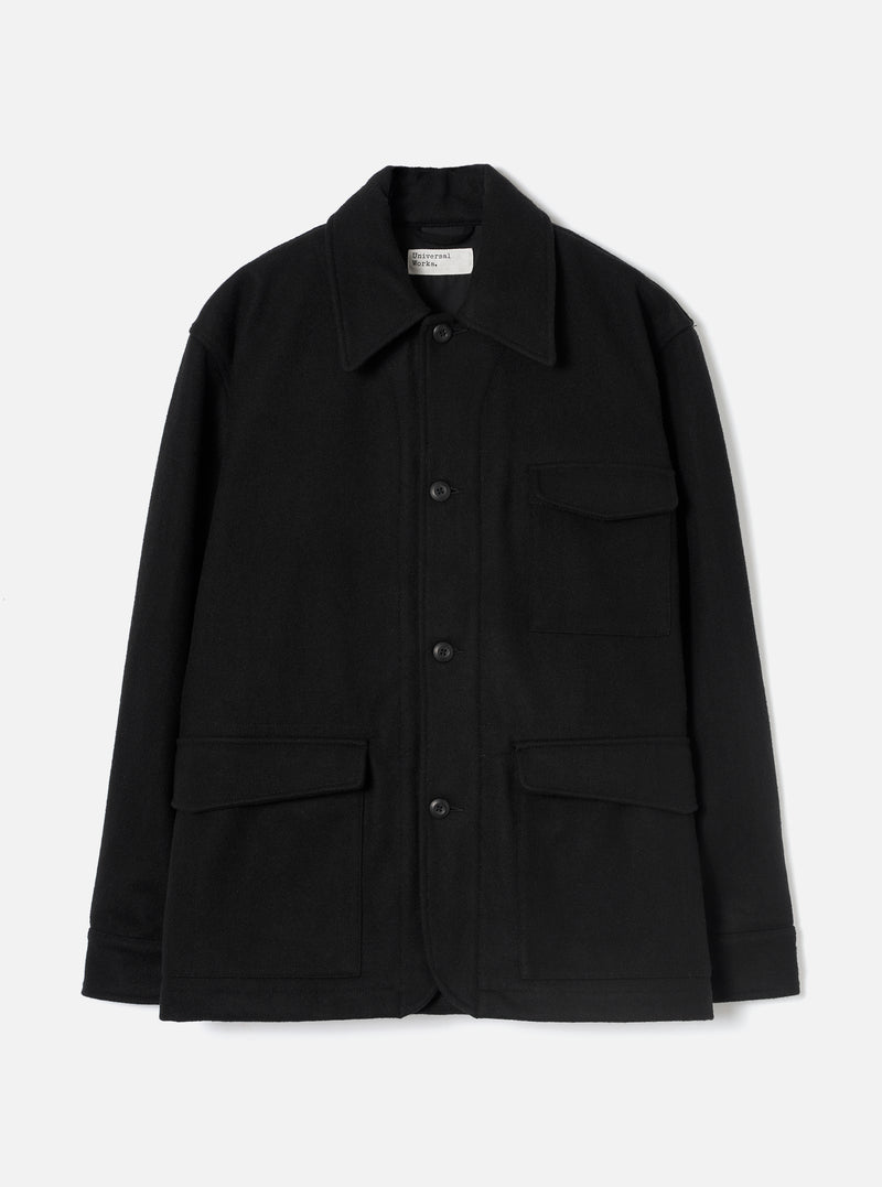Black wool-texture Mowbray heavy duty jacket laid flat on a light grey studio backdrop. Point collar, four-button front fastening, one chest patch pocket with flap, two large lower flap pockets, and long sleeves with plain cuffs. Universal Works woven label visible at the collar.