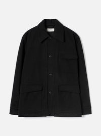 Black wool-texture Mowbray heavy duty jacket laid flat on a light grey studio backdrop. Point collar, four-button front fastening, one chest patch pocket with flap, two large lower flap pockets, and long sleeves with plain cuffs. Universal Works woven label visible at the collar.