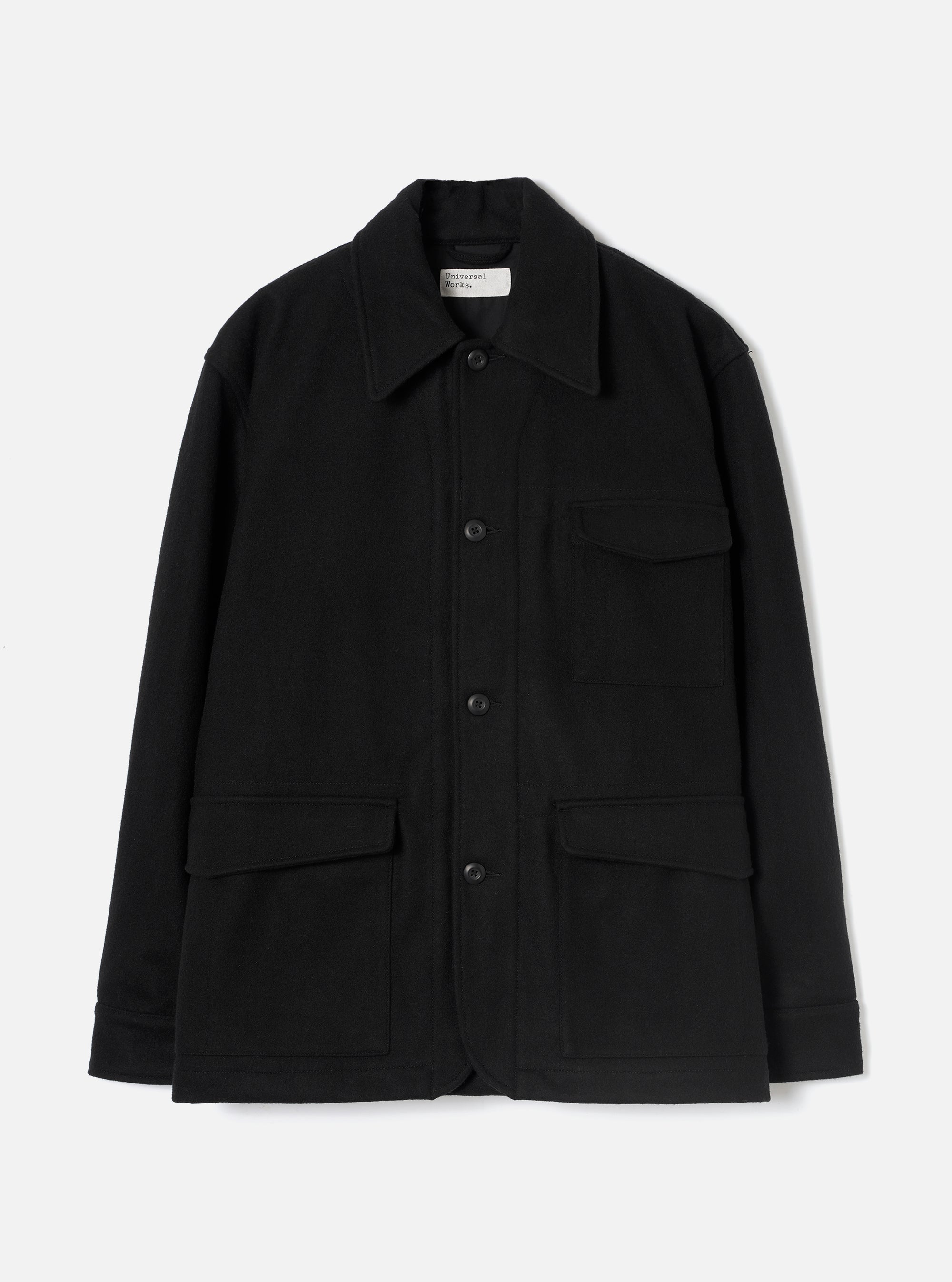 Black wool-texture Mowbray heavy duty jacket laid flat on a light grey studio backdrop. Point collar, four-button front fastening, one chest patch pocket with flap, two large lower flap pockets, and long sleeves with plain cuffs. Universal Works woven label visible at the collar.