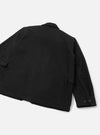 Black heavy duty jacket laid flat on a light grey surface, shot from the rear. A yoke seam runs across the upper back. Two flap pockets sit at the hip. The right sleeve is extended, showing a buttoned cuff with two dark buttons. The smooth, structured fabric has a matte finish.