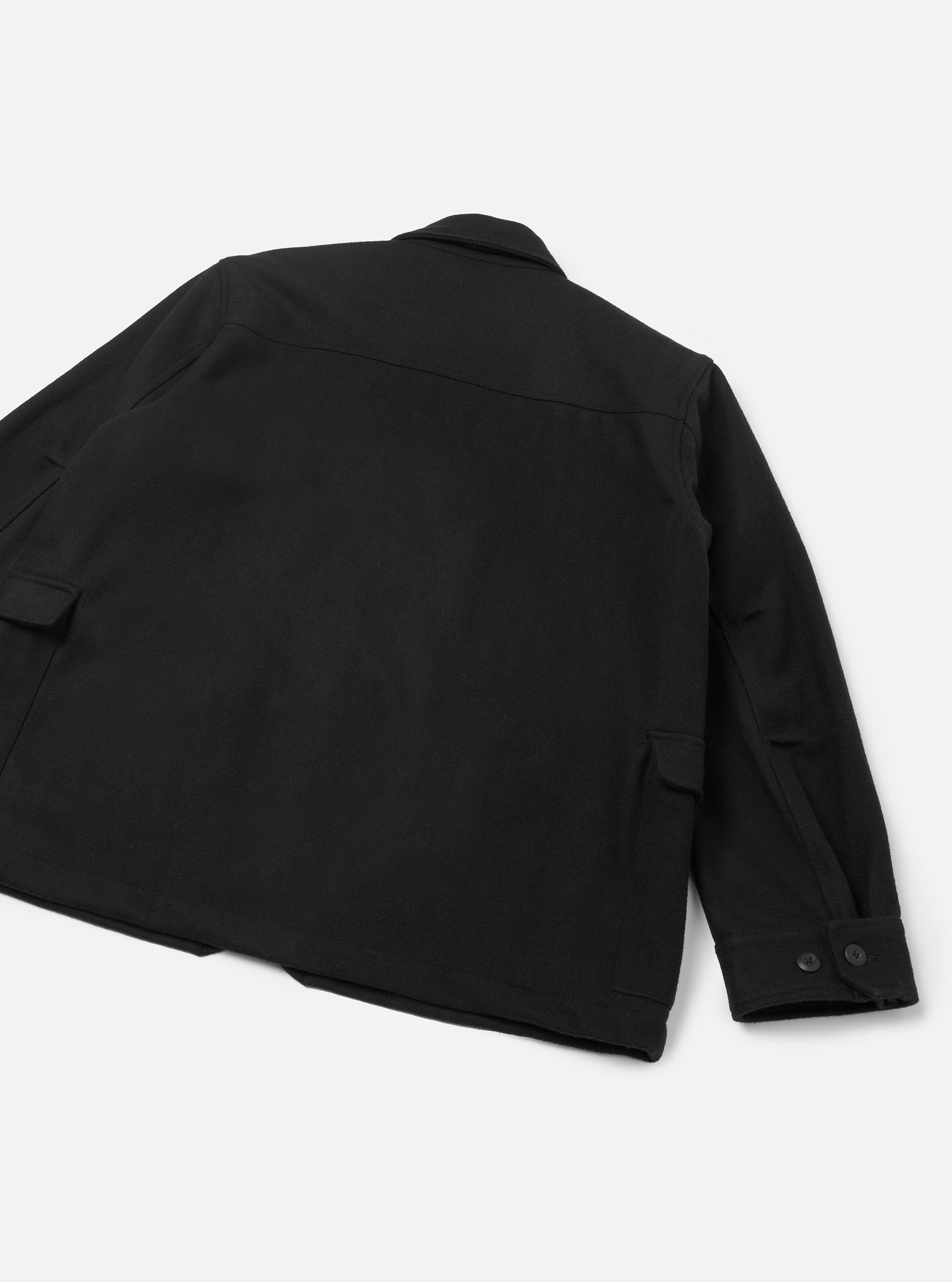 Black heavy duty jacket laid flat on a light grey surface, shot from the rear. A yoke seam runs across the upper back. Two flap pockets sit at the hip. The right sleeve is extended, showing a buttoned cuff with two dark buttons. The smooth, structured fabric has a matte finish.