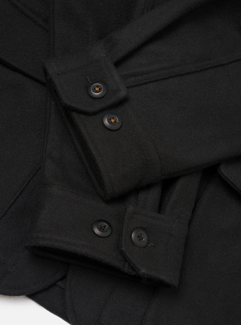 Close-up detail of the black Mowbray heavy duty jacket laid flat on a white surface. The shot focuses on the button placket and cuffs, showing dark four-hole buttons on a structured front placket and buttoned barrel cuffs. The fabric is a dense, matte wool-blend with a lightly brushed texture.