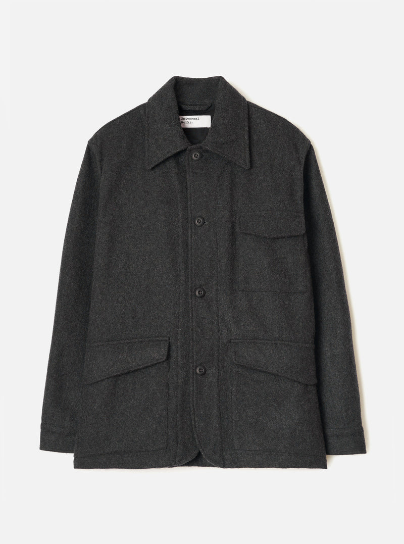 Grey marl wool chore jacket laid flat on a light grey seamless backdrop. Point collar with a Universal Works woven label at the neck. Four button front placket with dark buttons. Two chest patch pockets and two large flap pockets at the hem. Long sleeves with plain cuffs. Brushed, lightly flecked wool fabric throughout.