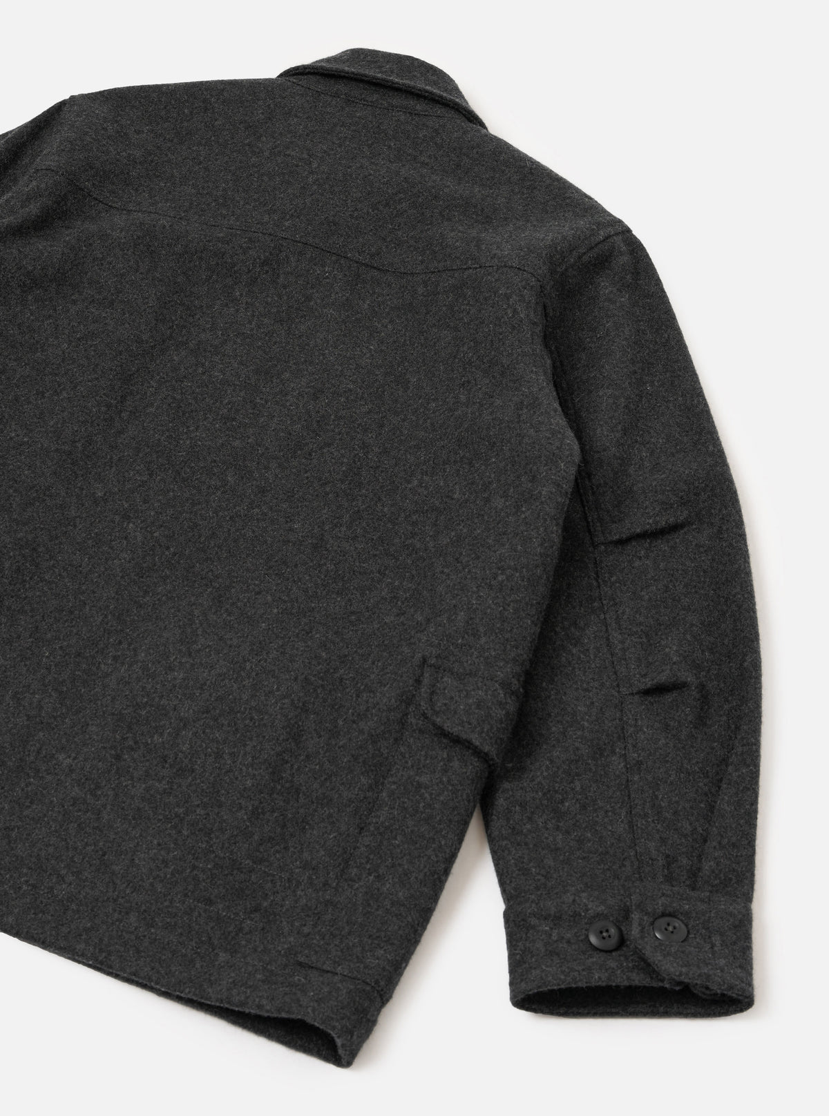 Flat lay of the grey marl heavy duty jacket on a white surface, shot from above at an angle showing the back and right sleeve. The melange wool fabric has a dense, felted texture. A yoke seam runs across the upper back. The cuff features two dark buttons, and a welt pocket is visible on the sleeve.
