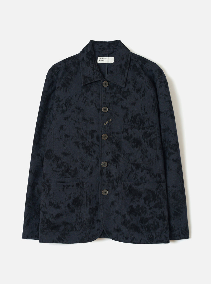 Black wool-mix chore jacket with an abstract inkblot jacquard pattern. Point collar, five dark horn-effect buttons down the centre front, a chest welt pocket and two large patch pockets at the hem. Full-length sleeves with plain cuffs. Universal Works label visible at the neck. Flat lay on a light grey studio backdrop.