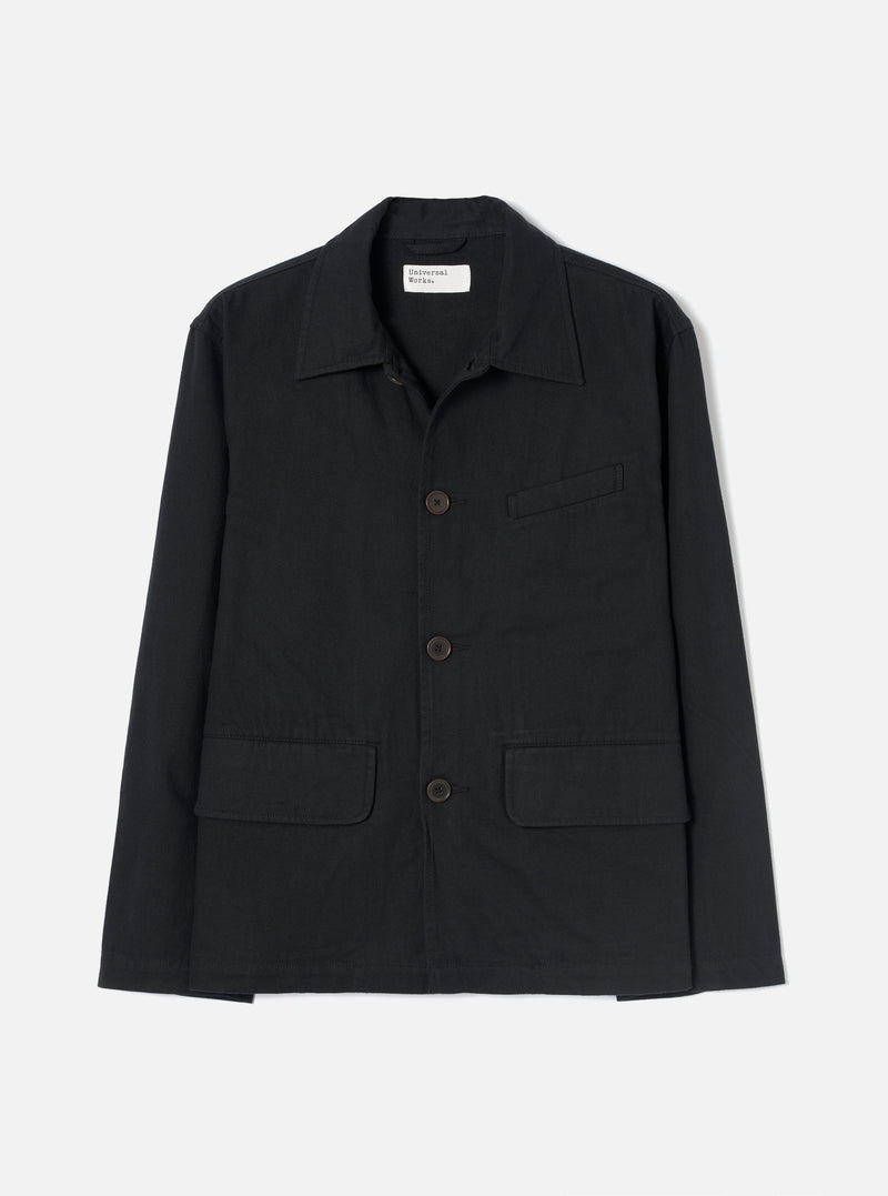 Black brushed moleskin chore jacket laid flat on a light grey seamless backdrop. Features a pointed shirt collar, three dark horn buttons down the centre front, two large patch pockets at the hem, one welt chest pocket, and long sleeves with plain cuffs. A Universal Works woven label is visible at the neck.