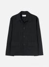 Black brushed moleskin chore jacket laid flat on a light grey seamless backdrop. Features a pointed shirt collar, three dark horn buttons down the centre front, two large patch pockets at the hem, one welt chest pocket, and long sleeves with plain cuffs. A Universal Works woven label is visible at the neck.