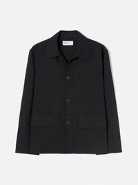 Black brushed moleskin chore jacket laid flat on a light grey seamless backdrop. Features a pointed shirt collar, three dark horn buttons down the centre front, two large patch pockets at the hem, one welt chest pocket, and long sleeves with plain cuffs. A Universal Works woven label is visible at the neck.