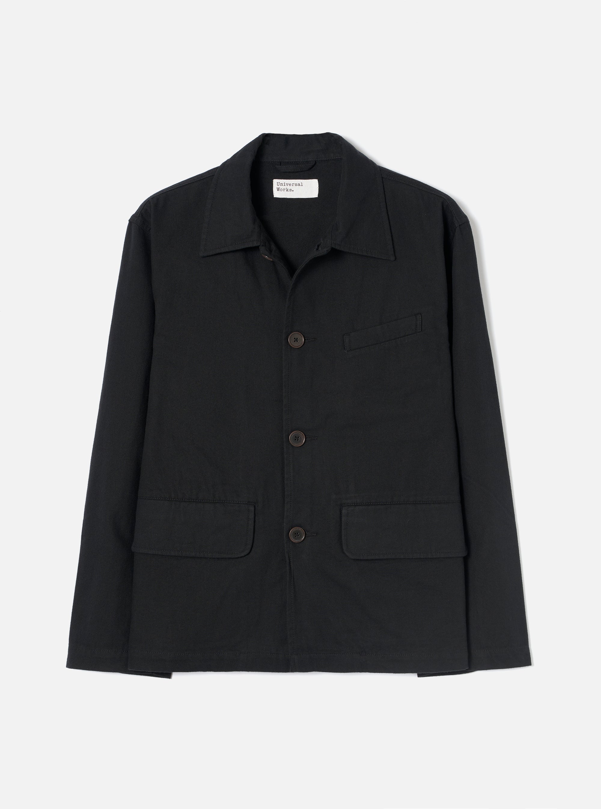 Black brushed moleskin chore jacket laid flat on a light grey seamless backdrop. Features a pointed shirt collar, three dark horn buttons down the centre front, two large patch pockets at the hem, one welt chest pocket, and long sleeves with plain cuffs. A Universal Works woven label is visible at the neck.
