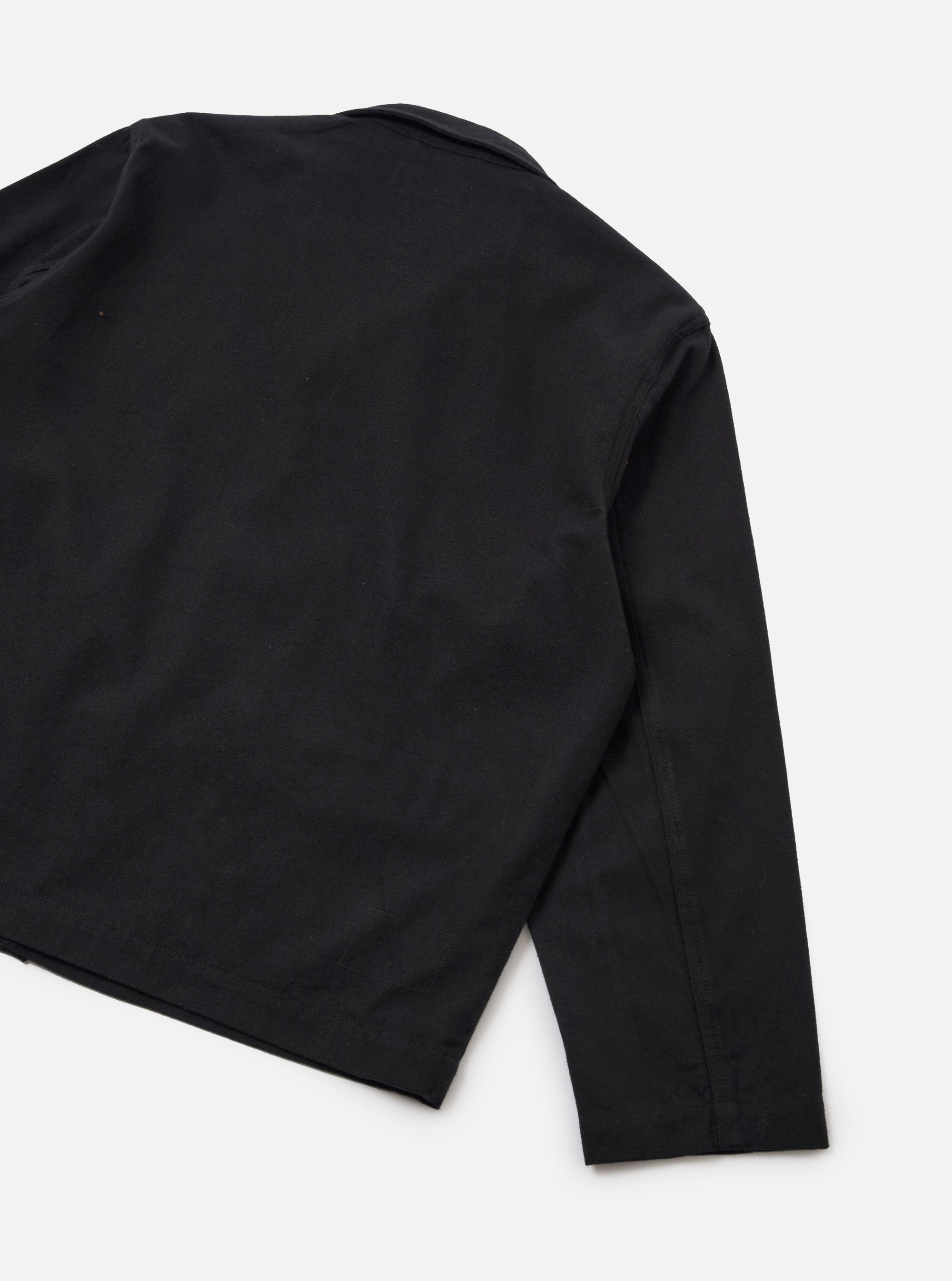 Rear detail flat lay of the black brushed moleskin Franck jacket on a plain white studio surface. The smooth, matte fabric has a subtle nap. Visible details include the back panel, a collar visible at the top, a set-in sleeve with a side seam, and a straight hemline.