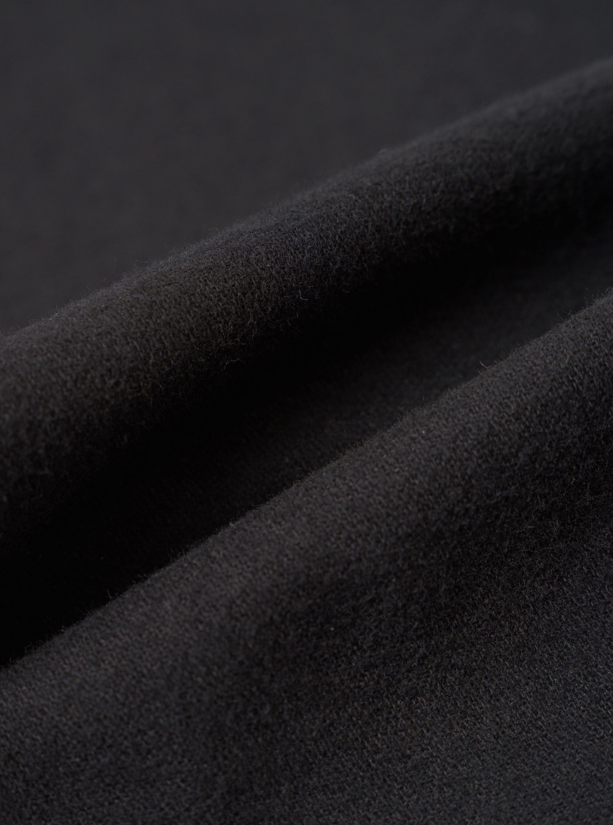 Close-up detail shot of the black brushed moleskin fabric used in the Franck Jacket. The macro image shows the dense, softly napped surface texture with gentle folds and ridges catching light to reveal the fabric's plush, fine-knit pile. Dark grey background.