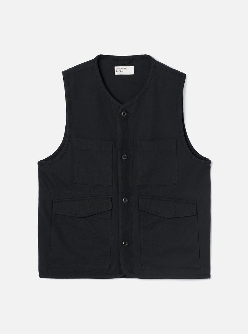 Black brushed moleskin gilet laid flat on a light grey studio backdrop. V-neckline with a Universal Works woven label at the back neck visible. Four button placket down the centre front with dark tonal buttons. Two chest patch pockets and two lower flap pockets. Sleeveless with armhole binding. Plain weave with a soft brushed texture.