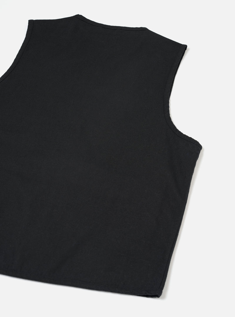 Black brushed moleskin gilet laid flat on a light grey surface, shot from above at a slight angle. The back of the garment is shown, revealing a crew neckline with topstitching, curved armholes with clean-finished edges, and a plain, unbroken back panel in smooth black fabric.