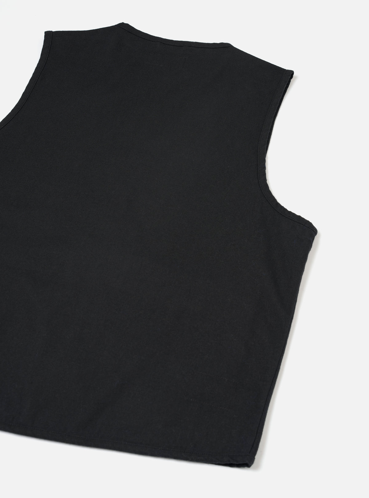 Black brushed moleskin gilet laid flat on a light grey surface, shot from above at a slight angle. The back of the garment is shown, revealing a crew neckline with topstitching, curved armholes with clean-finished edges, and a plain, unbroken back panel in smooth black fabric.